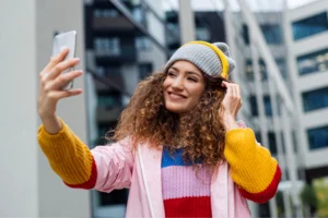 A smiling woman in a colorful sweater and gray beanie takes a selfie in an urban setting with modern buildings.