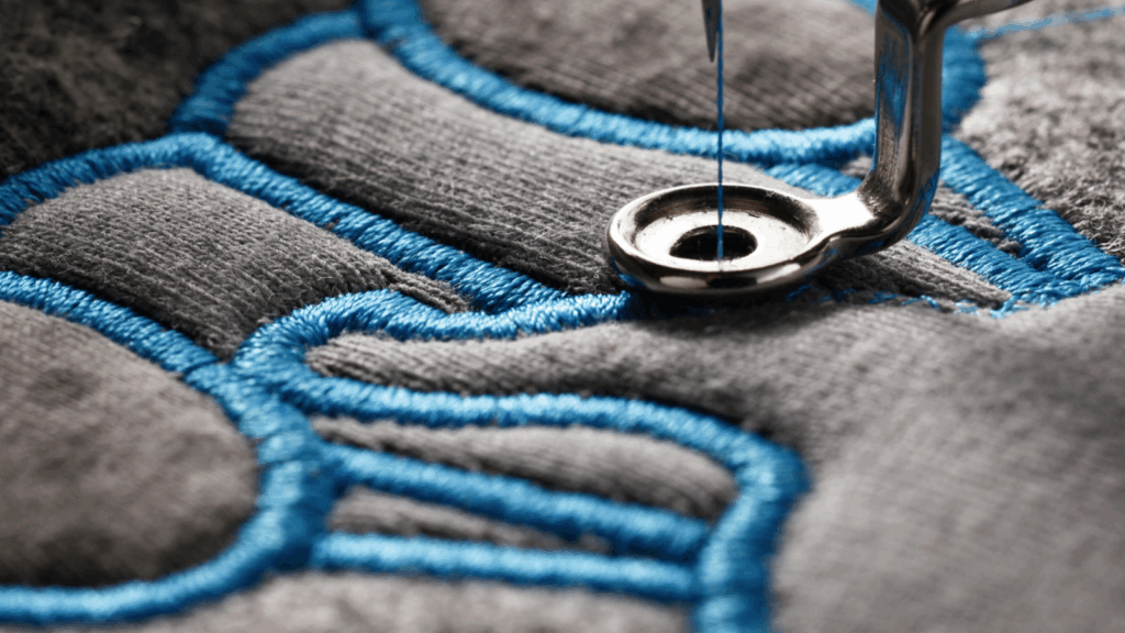 Close-up of a sewing machine needle stitching a detailed blue and grey embroidery logo on fabric.