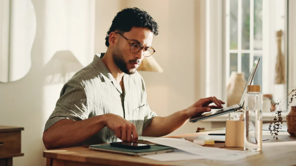 A man with glasses works at a desk, focusing on a tablet and a laptop, searching for TikTok Shop fees.