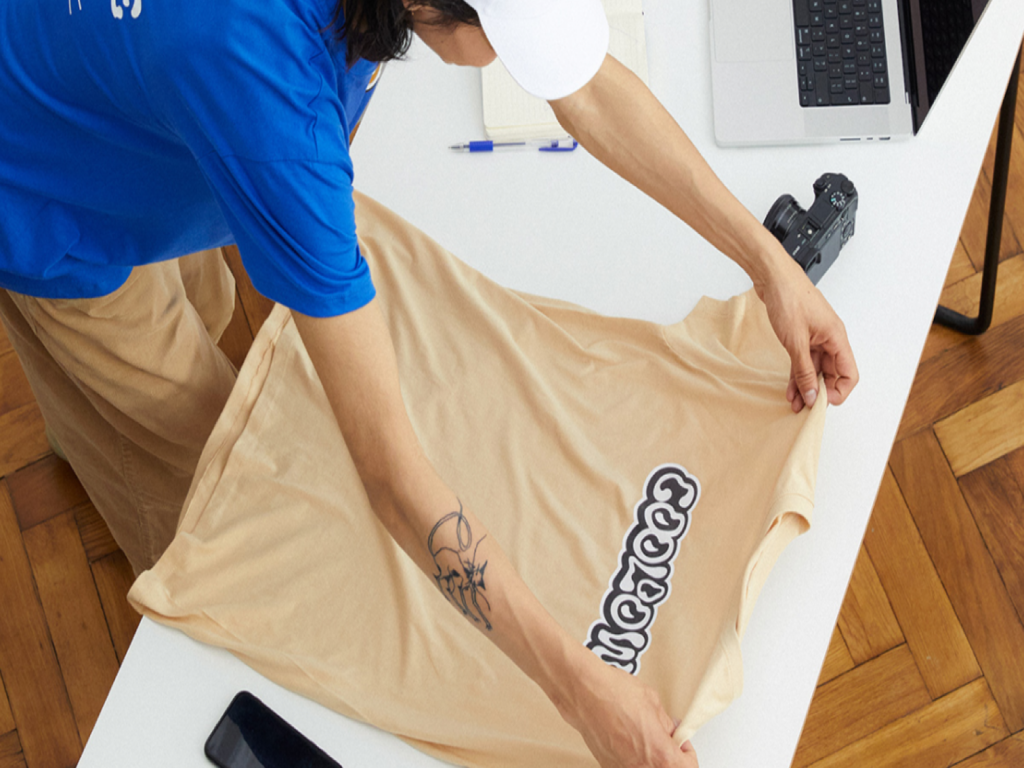 A person putting a custom t-shirt on top of a table, with a laptop and photo camera on the side of the t-shirt.