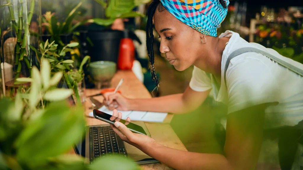 A woman in a colorful headscarf leans over plants, using a smartphone and laptop, taking notes.