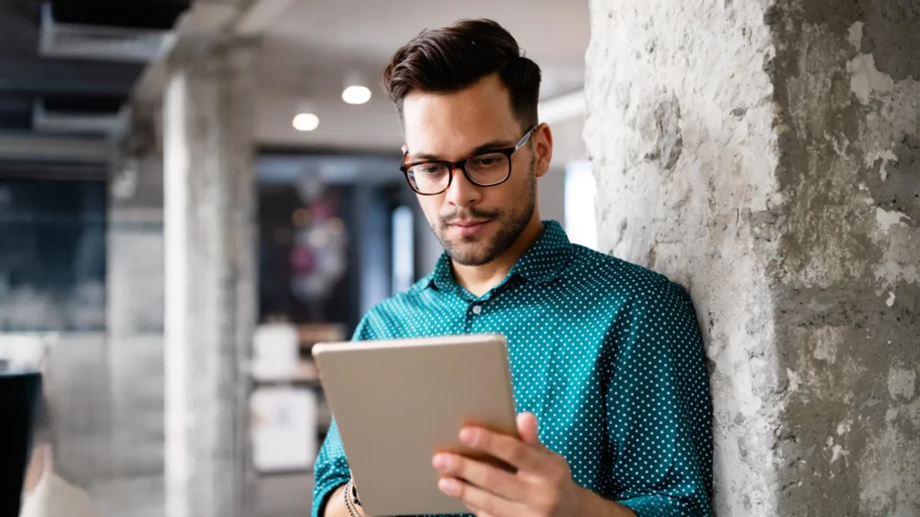 Etsy vs Shopify Which to choose for your business A man in glasses and a teal polka-dot shirt leans against a concrete pillar, reading a comparison of Shopify vs Etsy on a tablet.