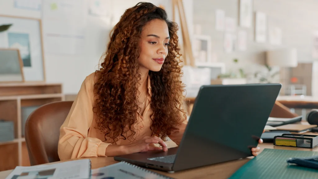 SEO and traffic generation A woman with curly hair, smiling, works intently on a laptop at a desk. The room is decorated with plants and artwork.