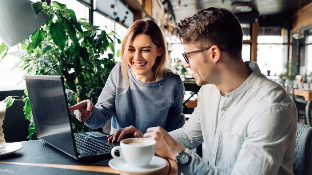 Scalability and international selling A smiling woman in a gray sweater points at a laptop screen, while a man in glasses and a white shirt sits beside her in a cafe.