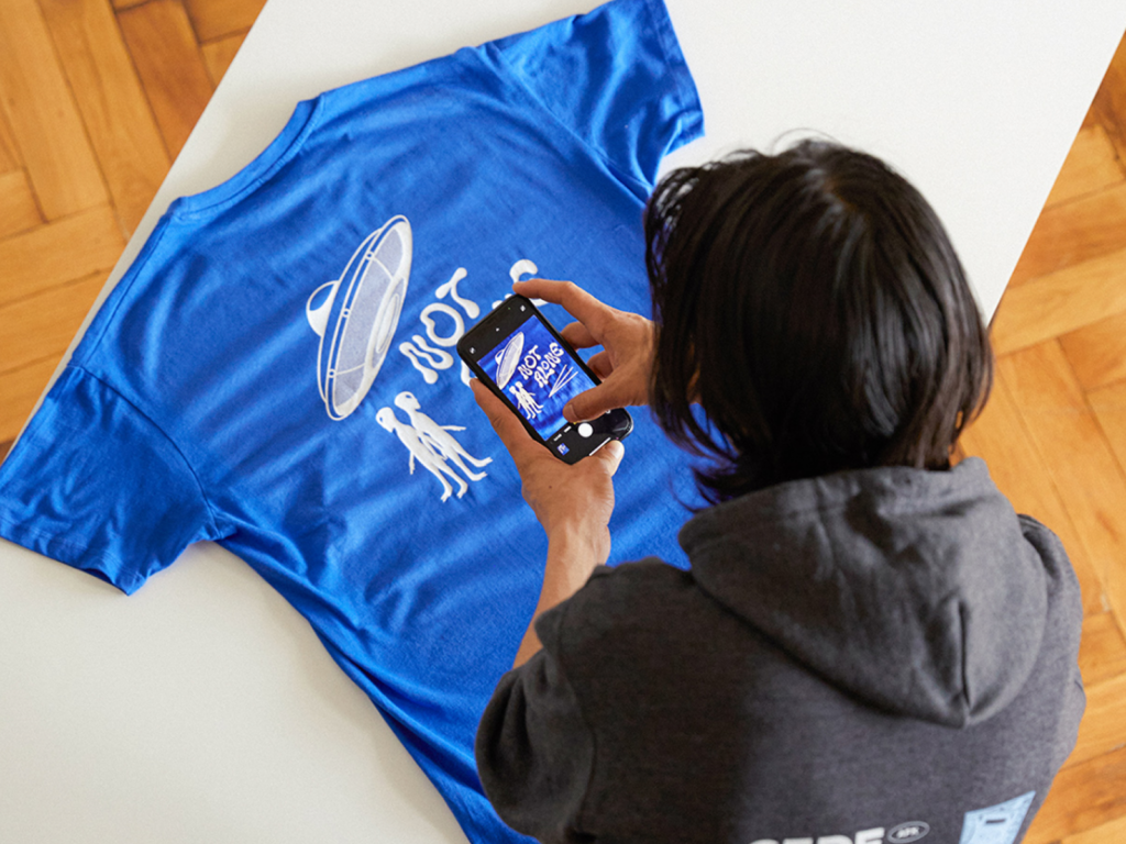 A person in a gray hoodie photographs a blue t-shirt featuring an alien and UFO design with "NOT ALONE" text.