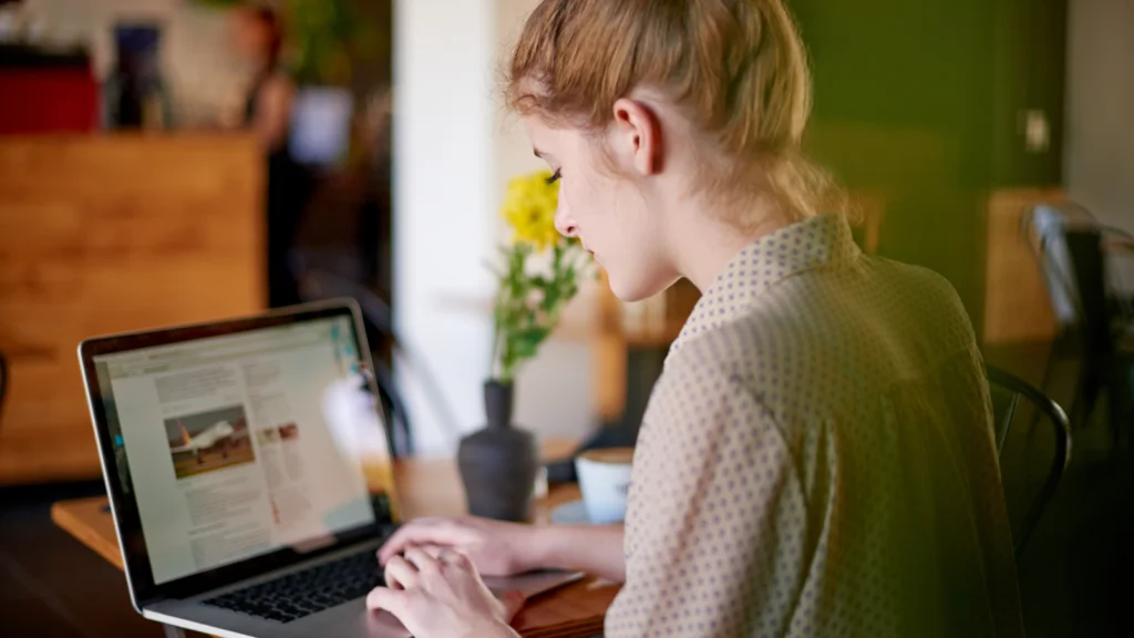 Storefront customization A woman with blonde hair works on a laptop, comparing Shopify vs Etsy, in a cozy cafe, focused on the screen. Yellow flowers in a vase nearby.