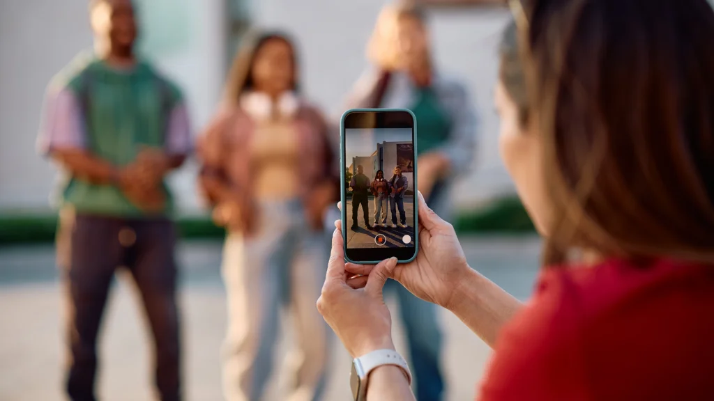 A person captures a video with a smartphone of three individuals standing outdoors, filming content for TikTok dropshipping.
