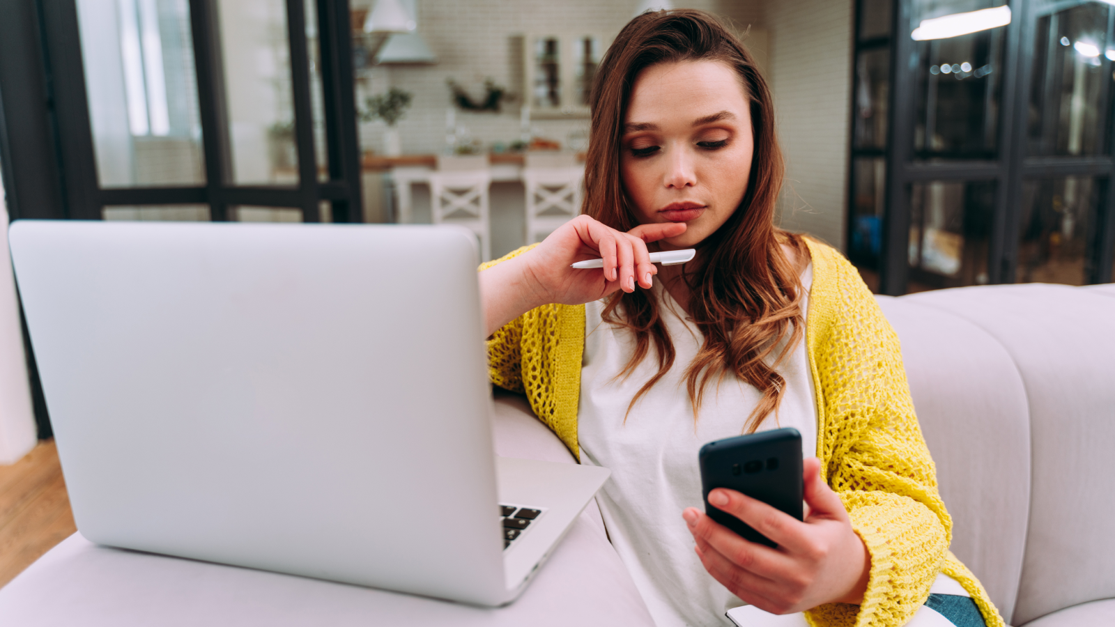 Advantages of Print on Demand for dropshipping A woman sitting on a couch in front of a laptop, while looking at a mobile phone in her hand.