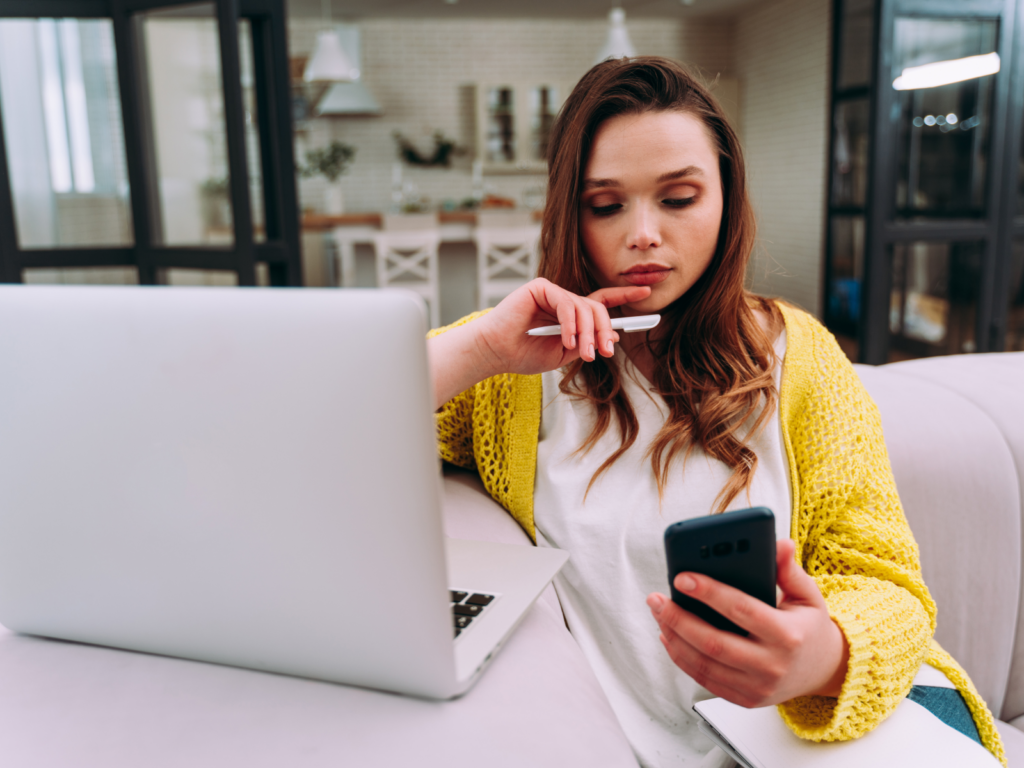 Become a Printify affiliate A woman sitting in front of a laptop, holding a pen in one hand and a mobile phone in the other.