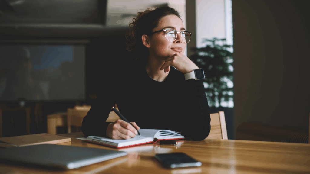 How much does the average shirt weigh A woman wearing glasses sits at a table, writing in a notebook with a pen.
