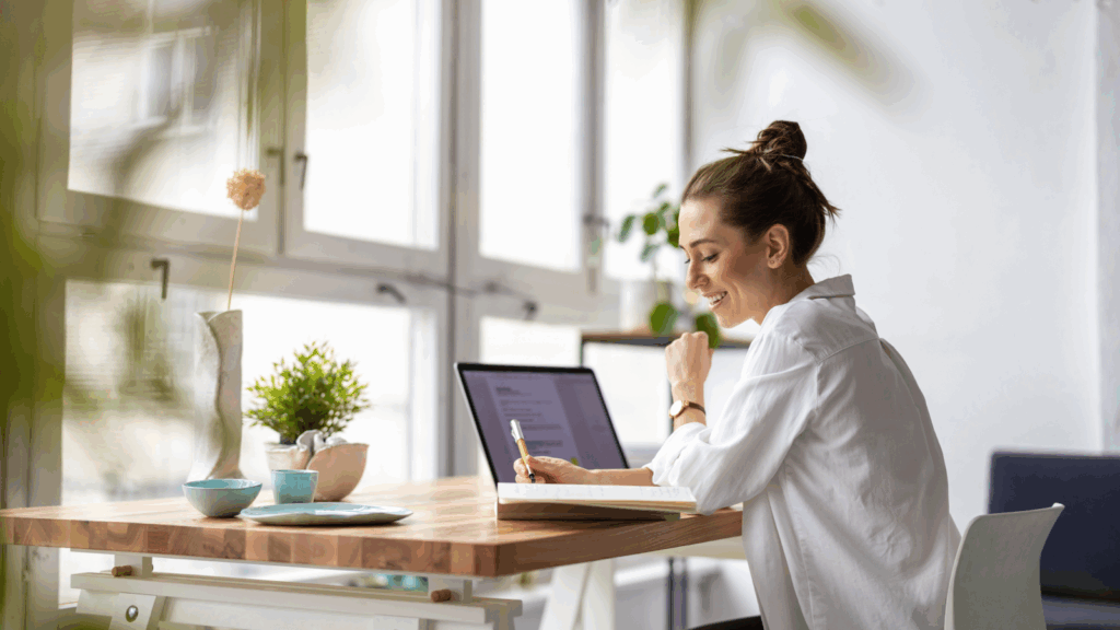 How to choose the right business for you A smiling woman in a white blouse sitting at a desk with a laptop on top of it, making notes.