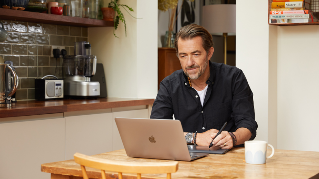 How to evaluate a POD companys production and supply chain A man in a blue shirt sitting at a kitchen table and using a laptop.
