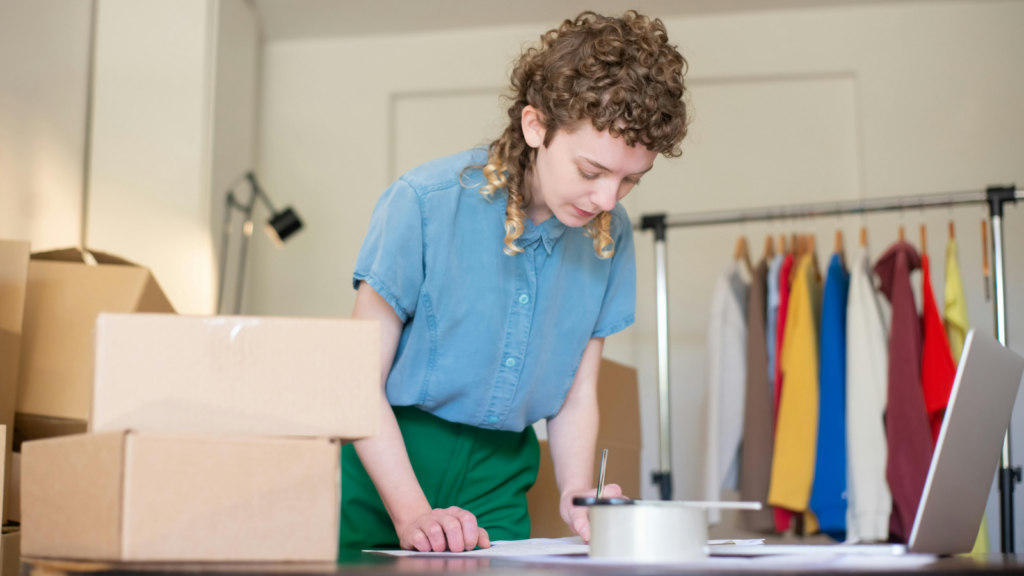 How to select the best products to sell on Shopify A woman standing in front of a table with delivery boxes on top, making notes.