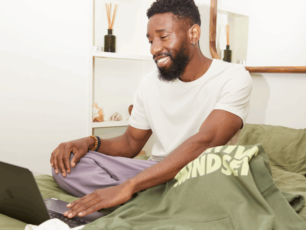 Make passive income with Printify A smiling man sitting on a couch with a laptop on his lap, and a green “Positive mindset” hoodie.