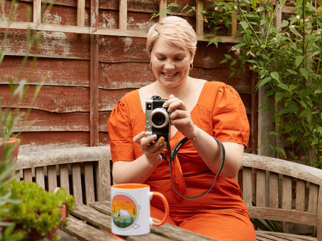 Print on Demand Passive income for artists A young woman in an orange dress taking a picture of a custom mug to earn passive income as an artist.