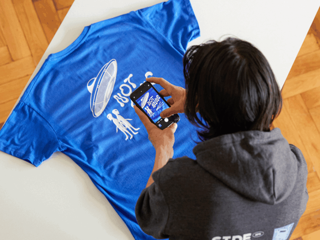Sell on Instagram with Printify A young man taking a picture of a blue t-shirt to sell on Instagram.