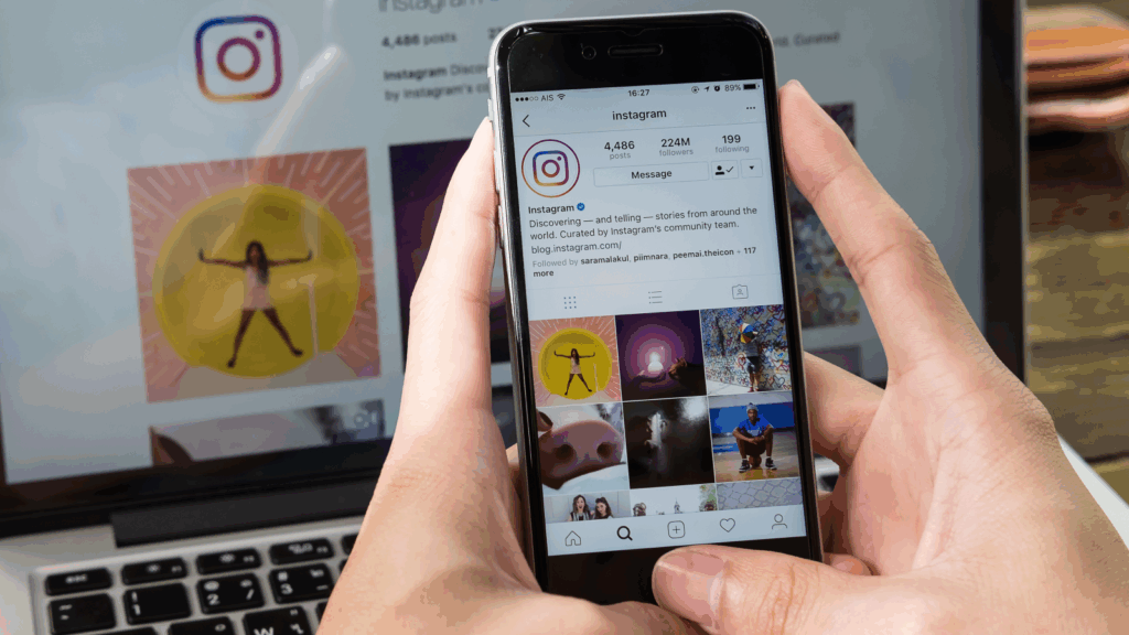 Set up your Instagram Shop A person holding a mobile phone with the Instagram app on the screen, with a blurred laptop in the background.