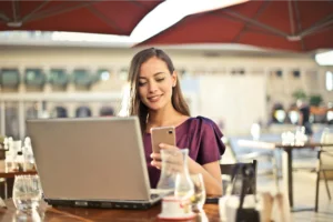 A smiling woman sitting in a cafe, using a mobile phone and laptop to do social media affiliate marketing.