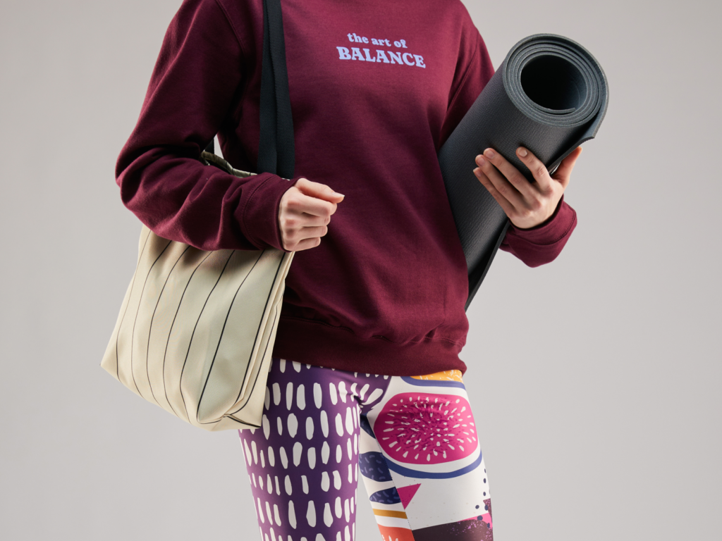 Sports and fitness products What to sell on Shopify A woman wearing custom sports apparel (purple hoodie, multi-colored yoga pants) and holding a yoga mat.