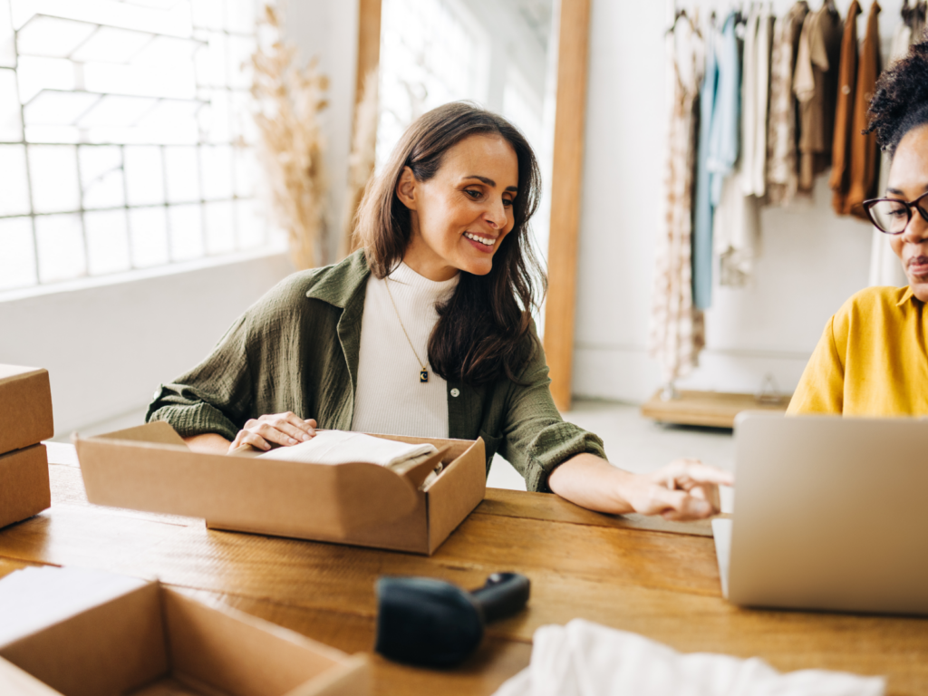 Start your eCommerce store with Printify A woman preparing a delivery box for shipment while conversing with another woman.