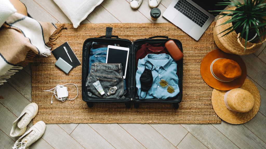Travel accessories What to sell on Shopify A travel bag full of clothing surrounded by different travel accessories (shoes, hats, laptop, charger).