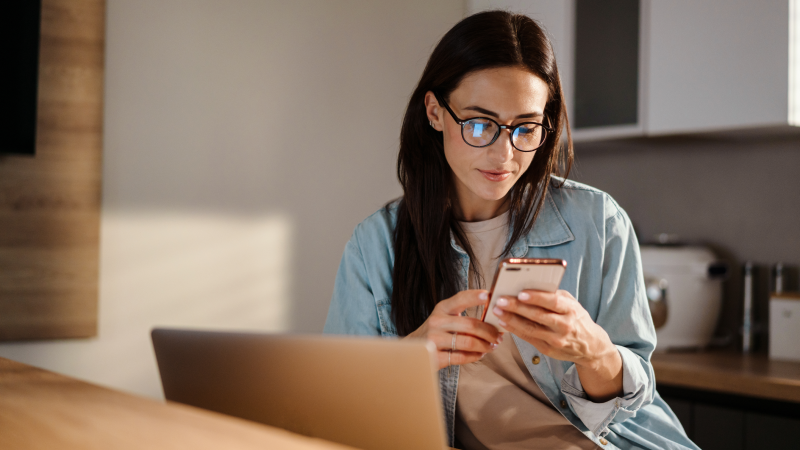 What is dropshipping A woman looking at her mobile phone while sitting at a desk in front of a laptop.