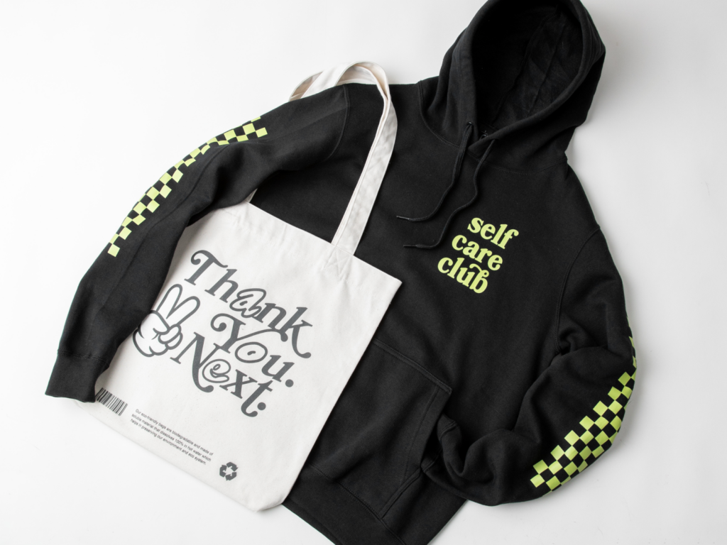 What makes a print on demand company eco friendly A custom black “Self-care club” hoodie next to a “Thank you. Next.” tote bag.