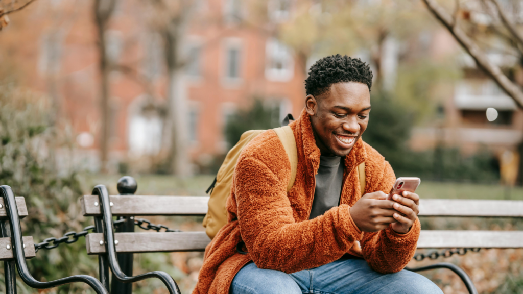 Why are social media and affiliate marketing a match A smiling man sitting on a park bench and using a mobile phone he’s holding in his hands.