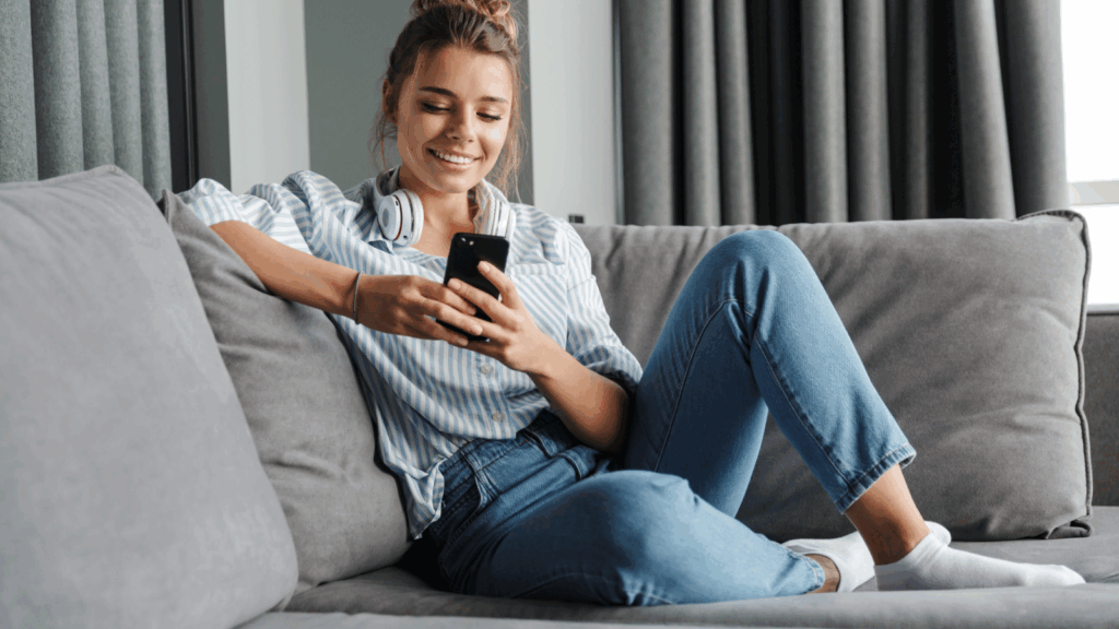 Benefits of using TikTok hashtags A smiling young woman is sitting on a couch and using a mobile phone to learn how to use TikTok hashtags.