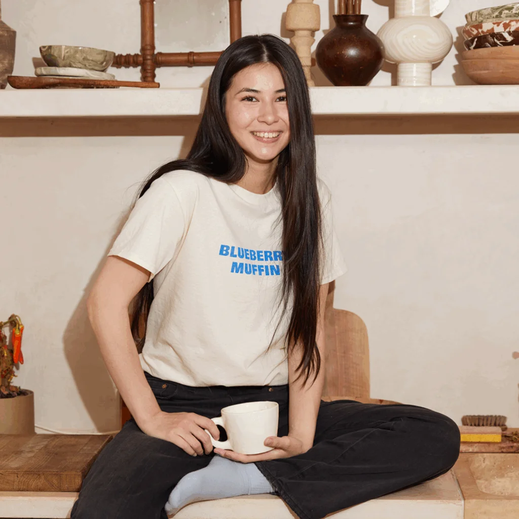 A smiling young woman wearing a light “Blueberry muffin” t-shirt.