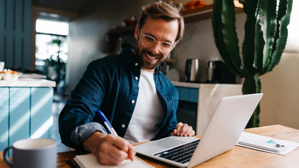 Creative Etsy shop name ideas for popular niches A smiling man with glasses writes in a notebook while using a laptop at a kitchen table. A large cactus is in the background.