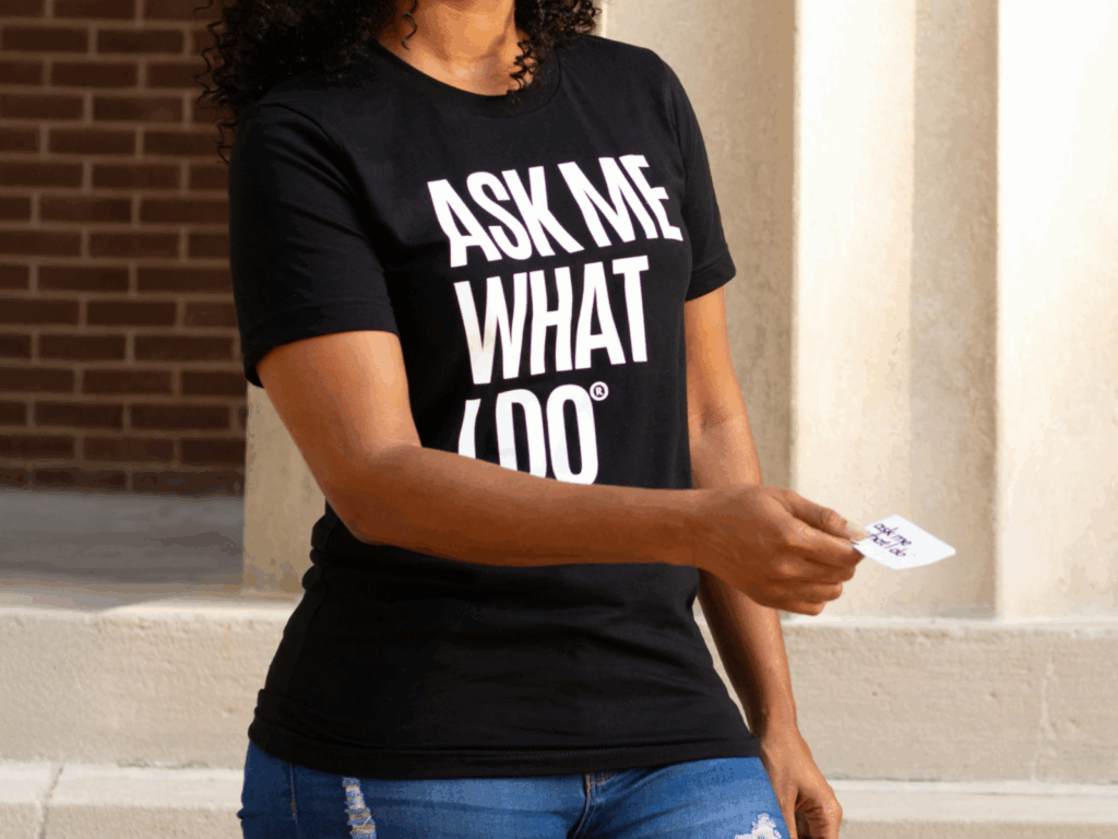 Design and order custom business cards with Printify A woman in a black “Ask me what I do” t-shirt is handing out a business card.