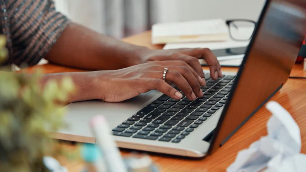Etsy shop name requirements and limitations Hands typing on a laptop keyboard on a wooden desk, surrounded by office items like papers and a plant.