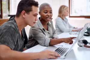 Two people are discussing Etsy shop names at a desk with laptops. The person on the right points at the screen.