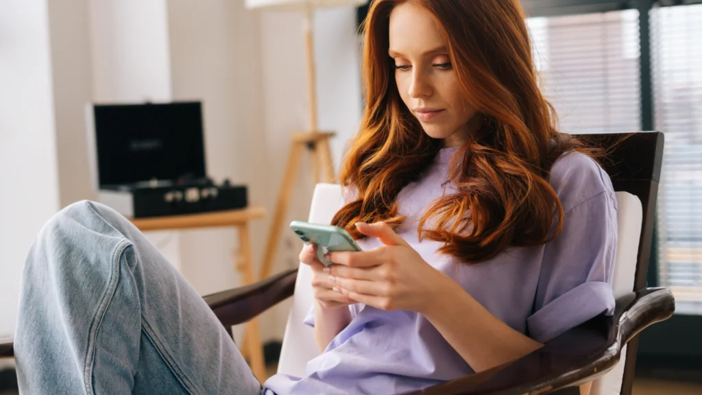Etsy shop naming mistakes that can hurt your brand A young woman with long red hair sits comfortably in a chair, focused on her smartphone. She wears a lavender shirt and jeans.