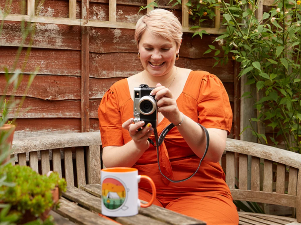 Got the Etsy store name List and sell products with Printify A smiling woman in an orange dress takes a picture of a colorful mug on the table, with greenery surrounding her.