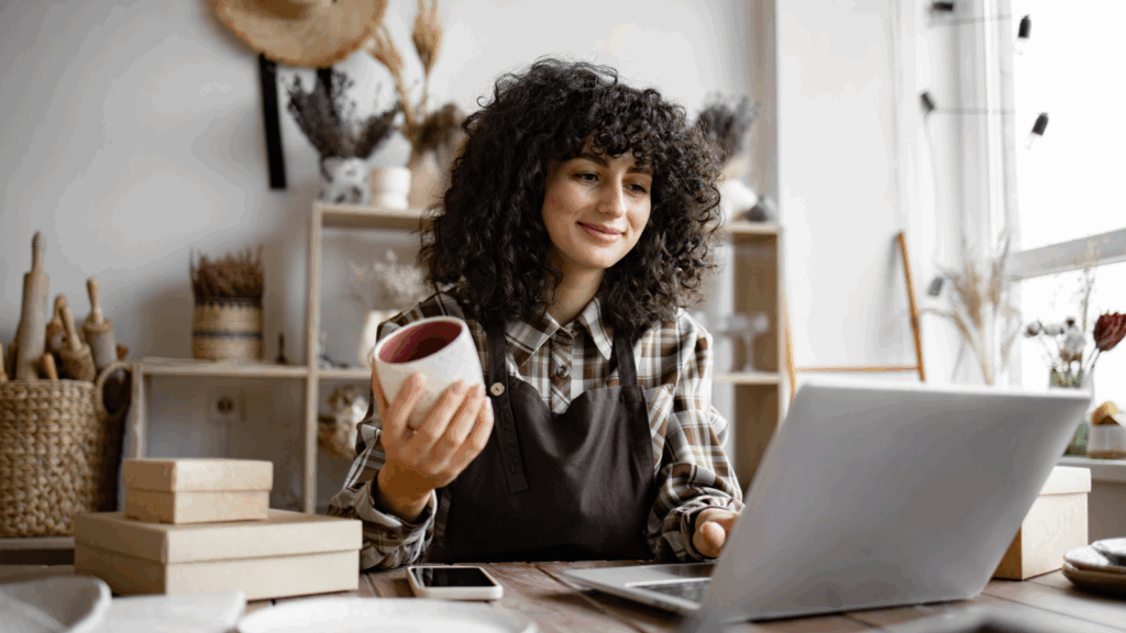 How do you sell contemporary art online A woman with curly hair sits at a table, focused on her laptop, ready to sell her art online.