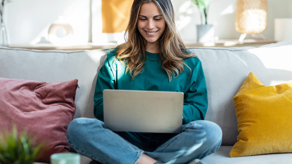 How to come up with Etsy shop name ideas A smiling woman in a green sweater sits cross-legged on a sofa, using a laptop to search for Etsy shop name ideas.