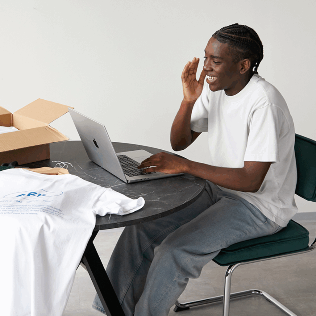 How to dropship for free with Printify A smiling young man is sitting at a table using a laptop. Custom t-shirts are placed near the laptop.