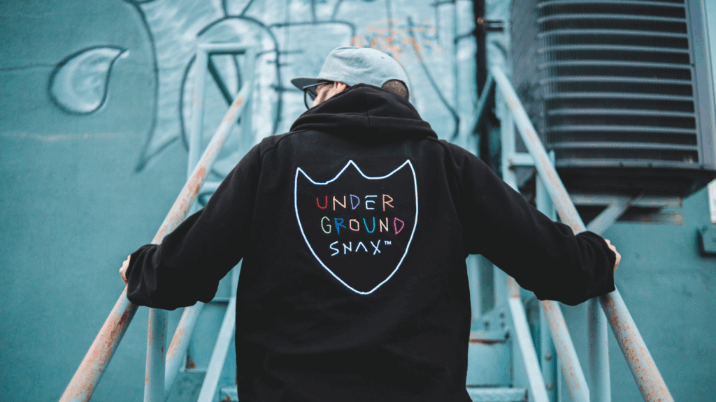 How to get started with custom embroidery on Printify A young man in a black hoodie with “Underground snax” embroidered on the back of the hoodie.