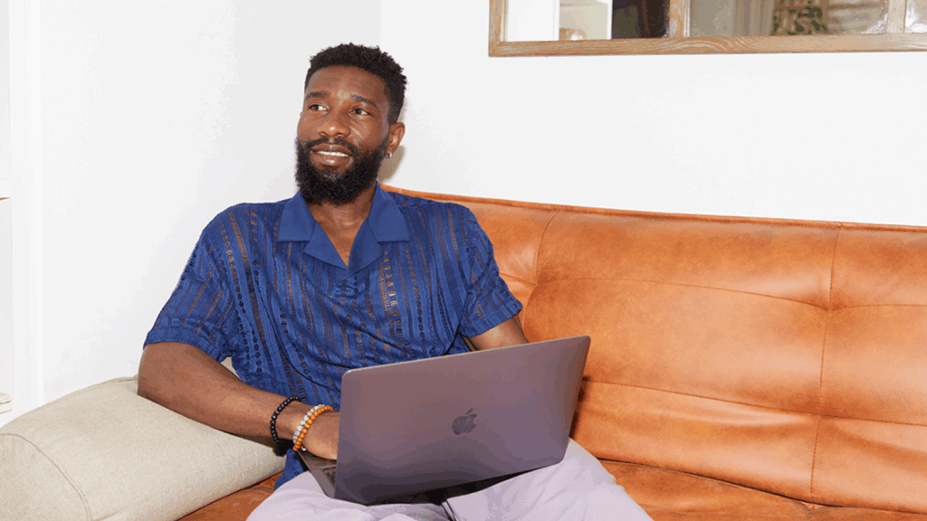 Print with print on demand A smiling man in a blue shirt is sitting on a couch and using a laptop.