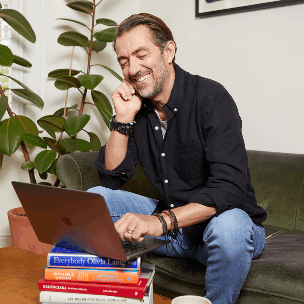 Print your canvas hassle free with Printify A smiling man using a laptop that’s standing on a stack of books.
