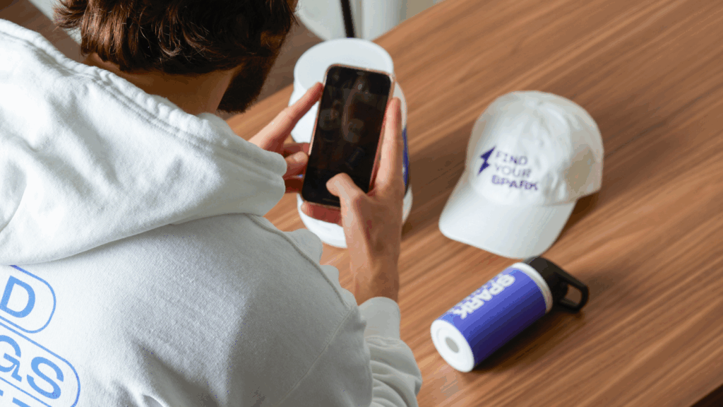 A man in a white hoodie and blue hat takes a photo with his phone, focusing intently on the screen.