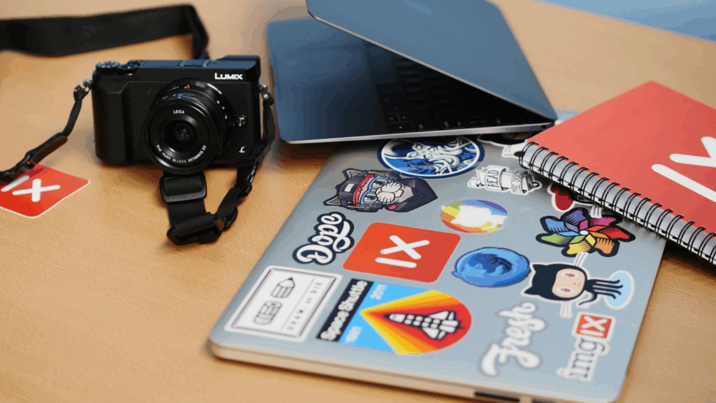 Stickers Etsy shop ideas A laptop, camera, stickers and various items arranged neatly on a wooden desk, showcasing a workspace setup.