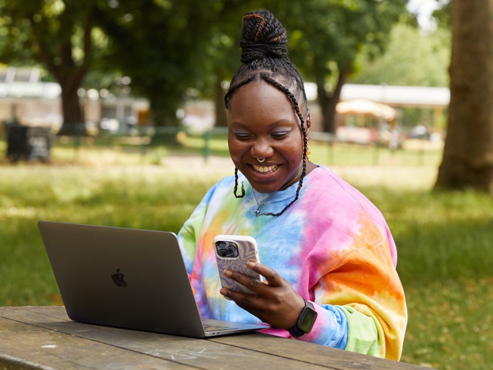 Tips for successfully selling customized products A smiling woman is looking at her phone while sitting at a bench in a park and using a laptop.