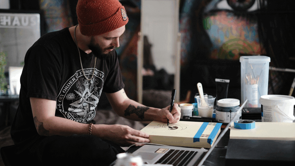 Tips on how to make a living as an artist A man wearing a beanie works intently on his laptop at a desk, focused on his task.