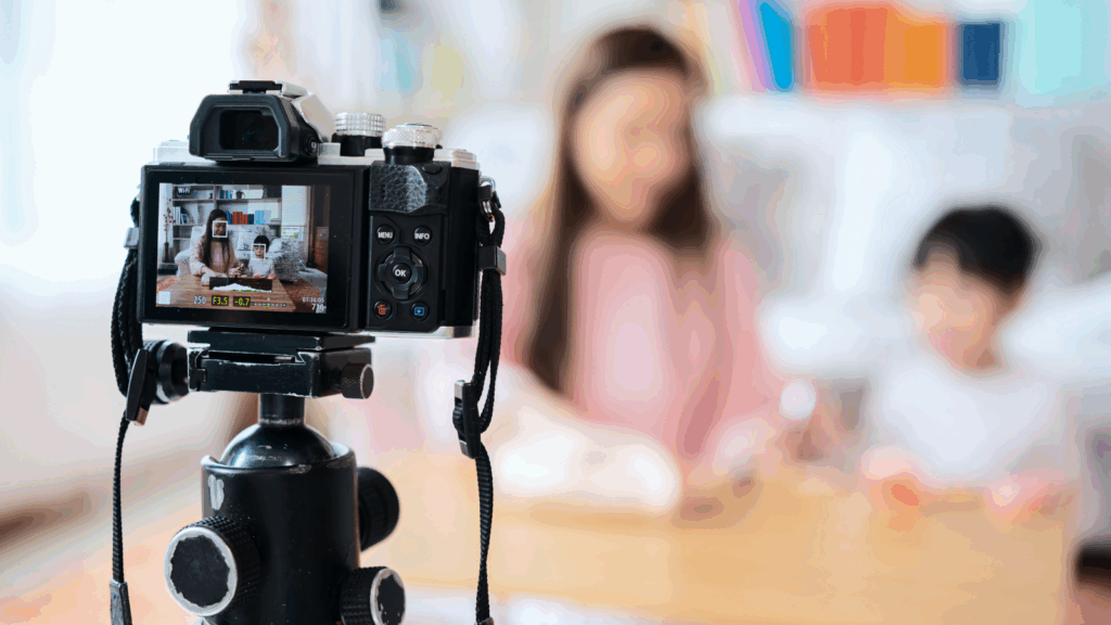 A camera in focus is recording with a blurred background of a woman and a child.