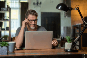 A man in glasses is sitting at a table and reading a blog, Big Cartel vs Shopify, on a laptop.