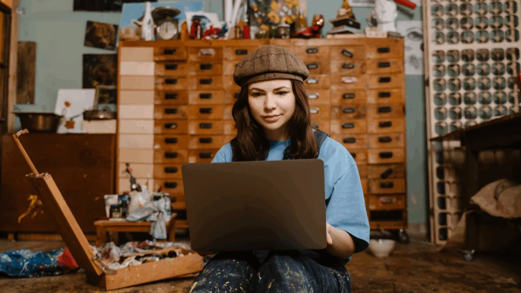 A woman painter is sitting on the floor with a laptop on her lap.