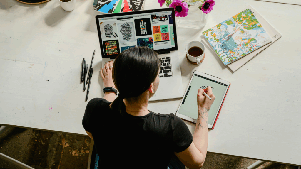 A woman is sitting at a desk, using a laptop and pad to create graphic design elements.